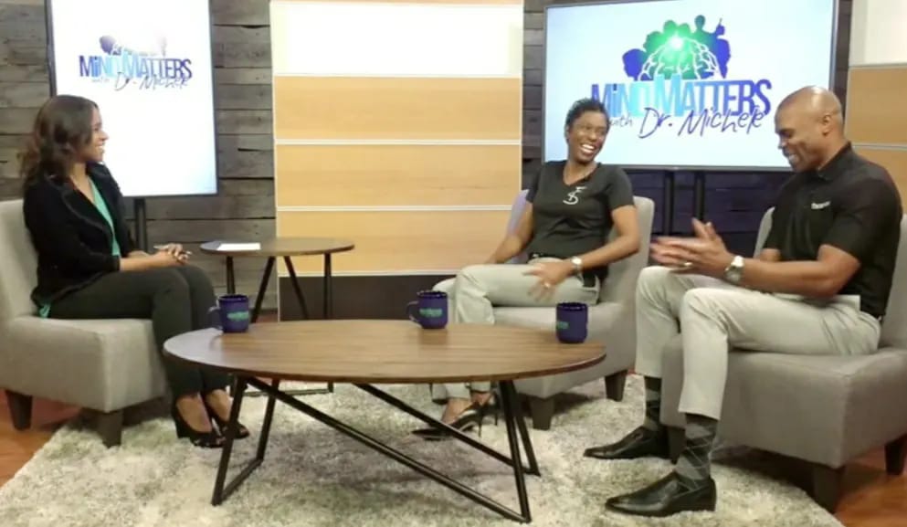 Three people seated in a studio interview setting with microphones and monitors displaying a logo.