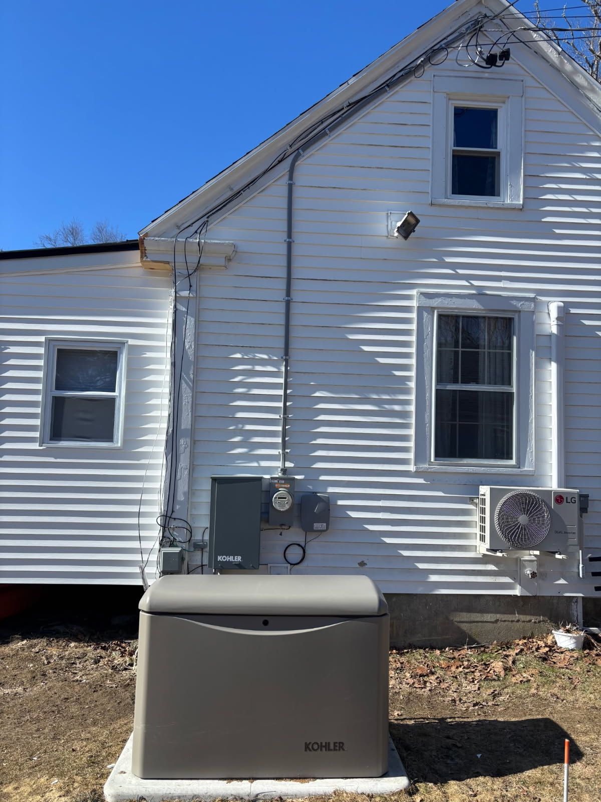 A white-sided house with an outdoor backup generator, a mounted electric meter, and a small HVAC unit on a sunny day.