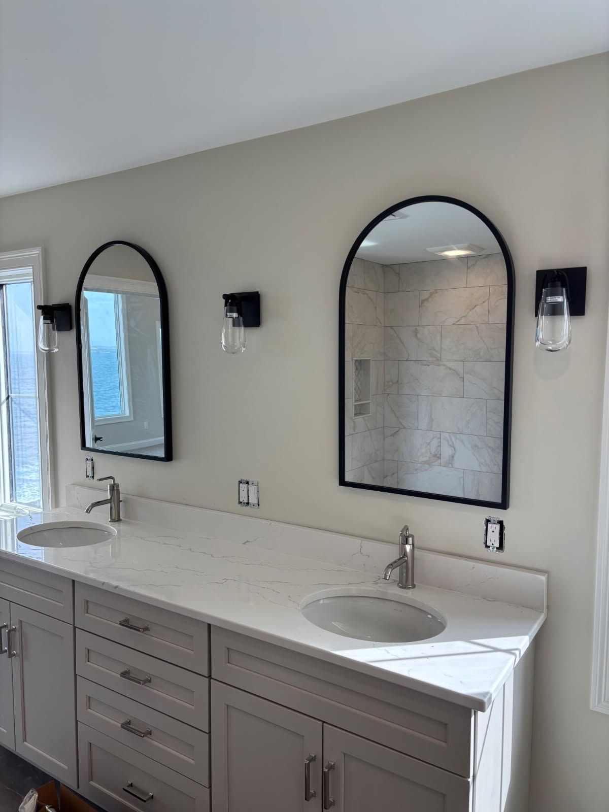 A double bathroom vanity with light gray cabinets, white marble countertops, and two arched black-framed mirrors.