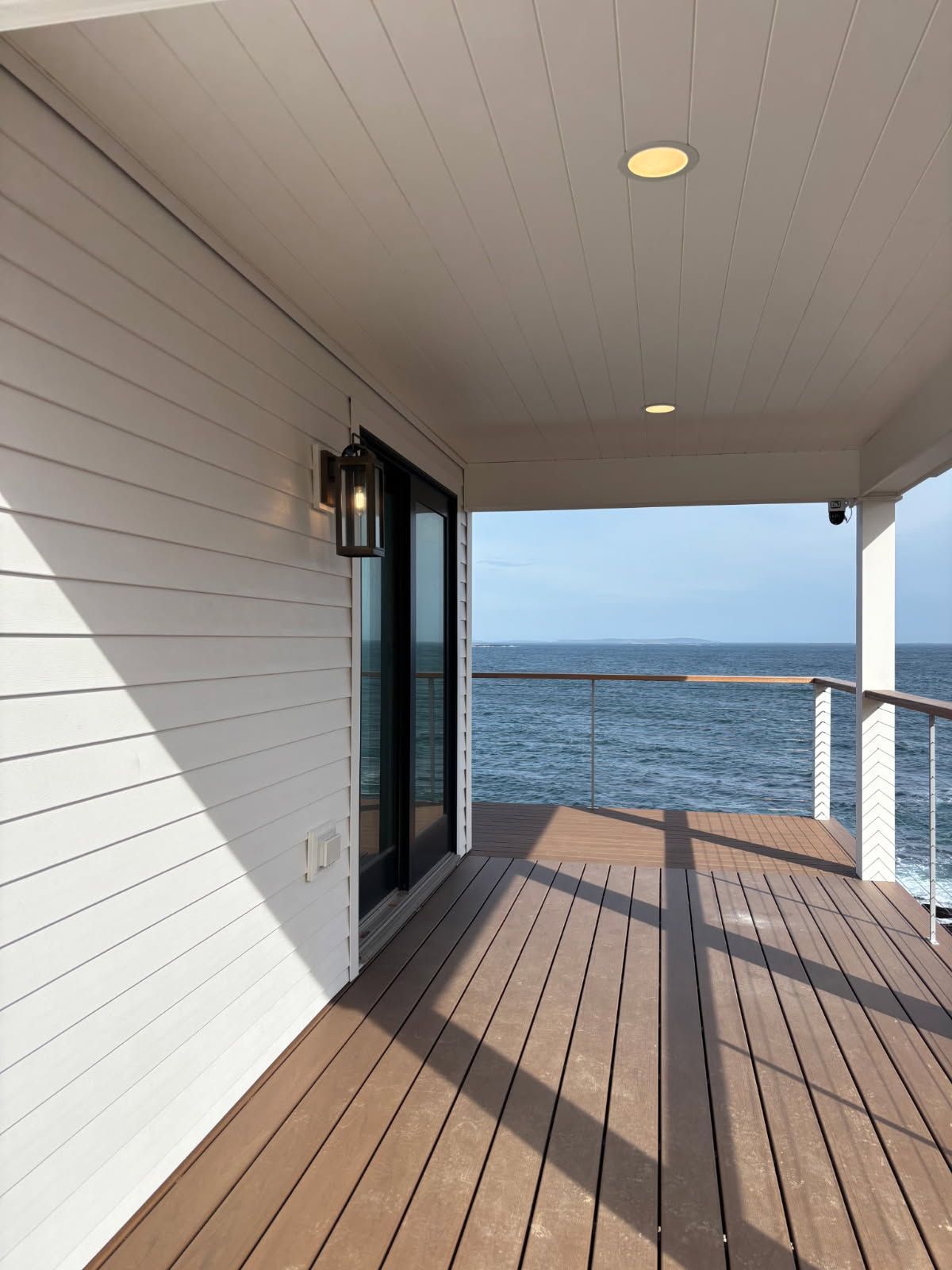 A bright deck with white siding and brown flooring, overlooking the ocean under a clear sky.