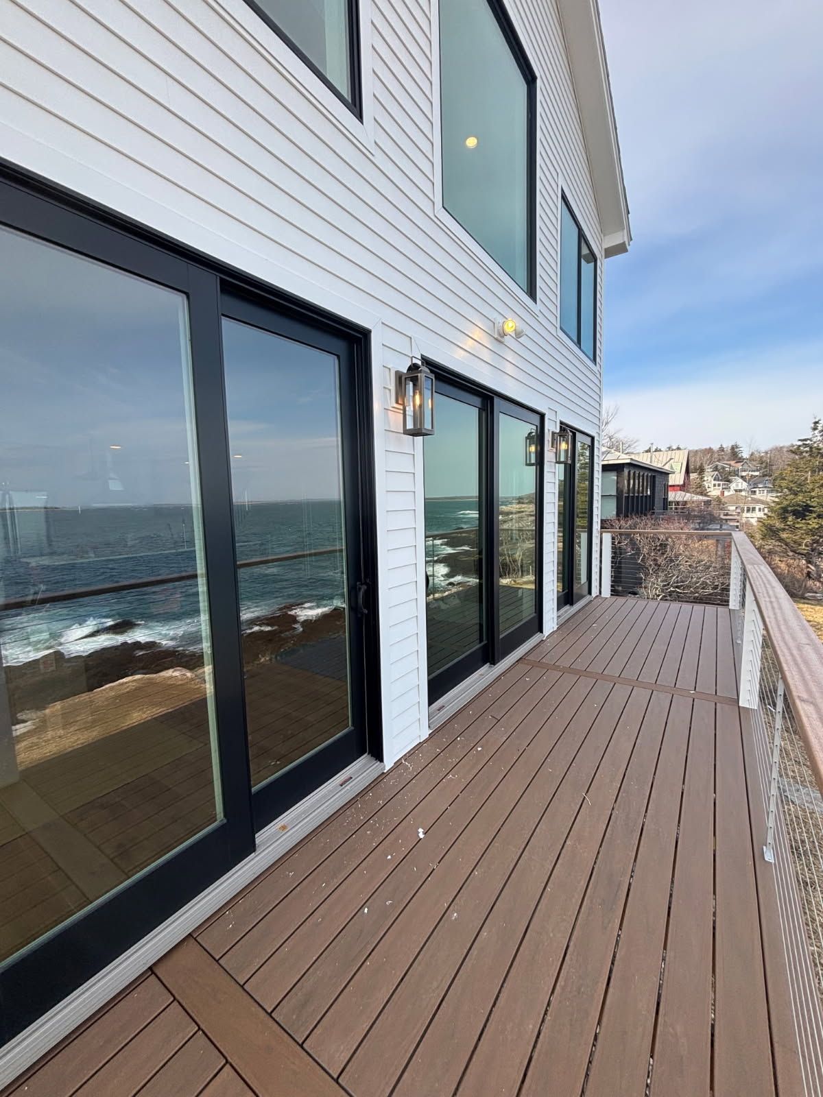 A wooden deck with a railing overlooks a rocky coastline and the ocean next to a white house with large glass doors.