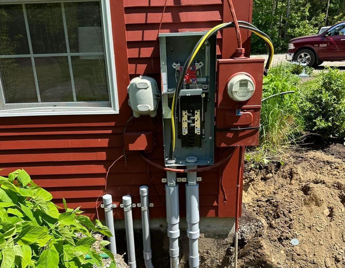 An open electrical service panel with conduits entering from the ground, mounted on the side of a red house.