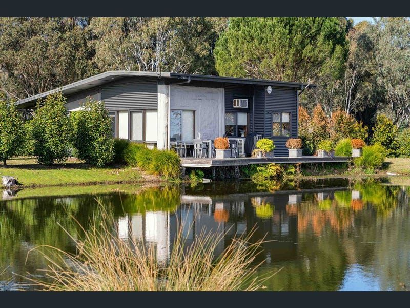 A modern house with dark siding sits by a reflective pond, surrounded by trees and a deck with potted plants.