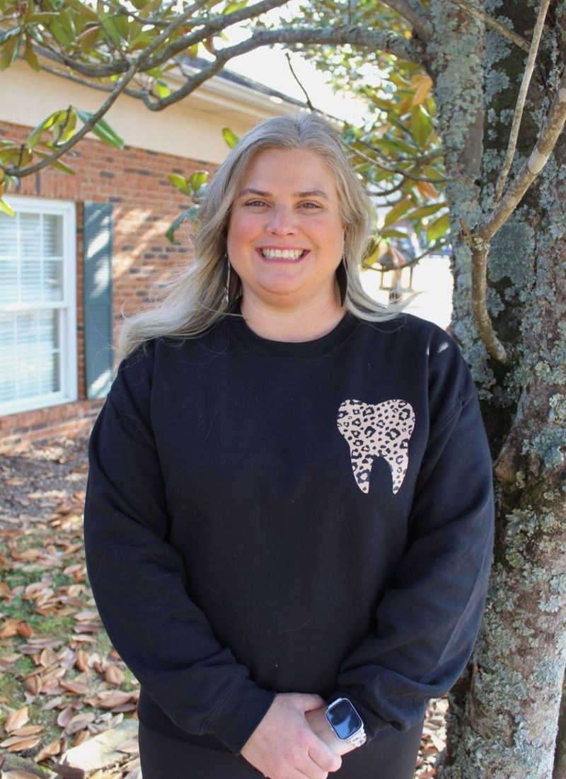 A woman wearing a black sweater with a tooth on it is standing in front of a tree.