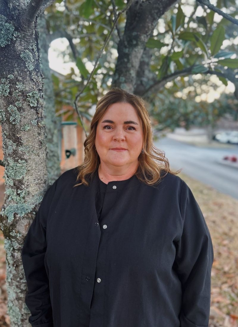 A woman in a black jacket is standing next to a tree.