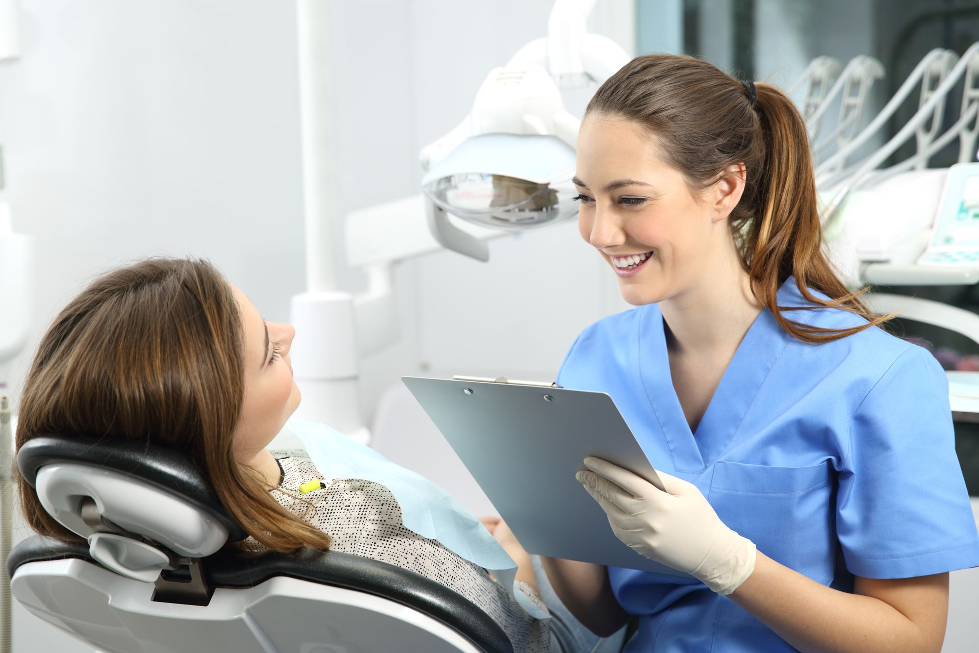 A dentist is talking to a patient in a dental chair while holding a clipboard.