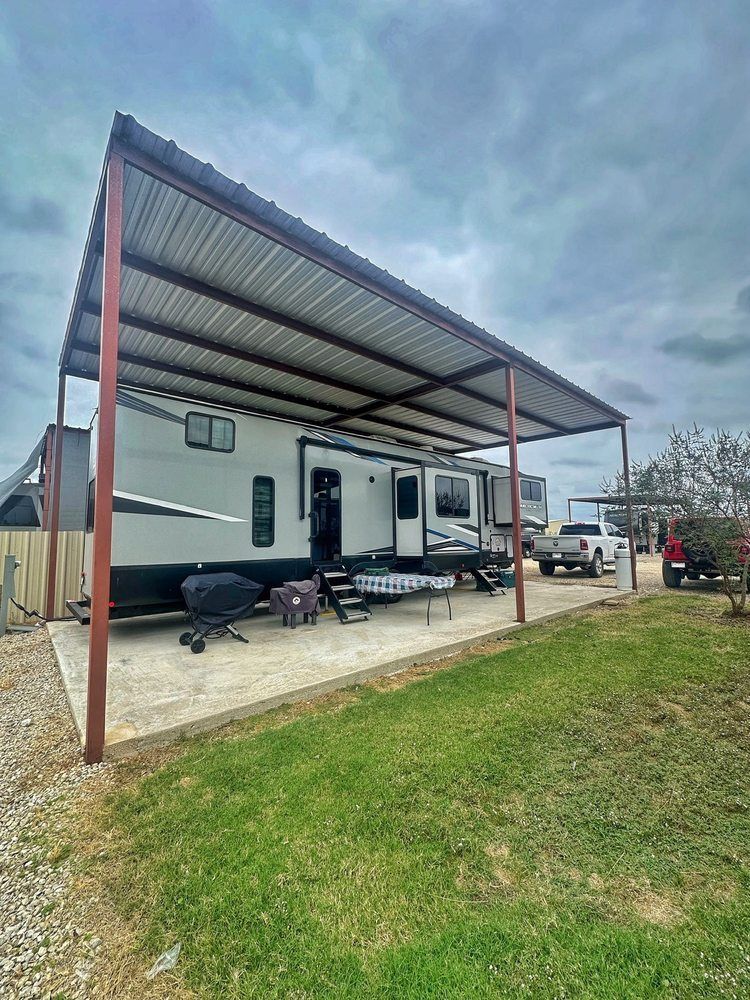 RV parked under a metal awning on a concrete pad with a grassy area in front.