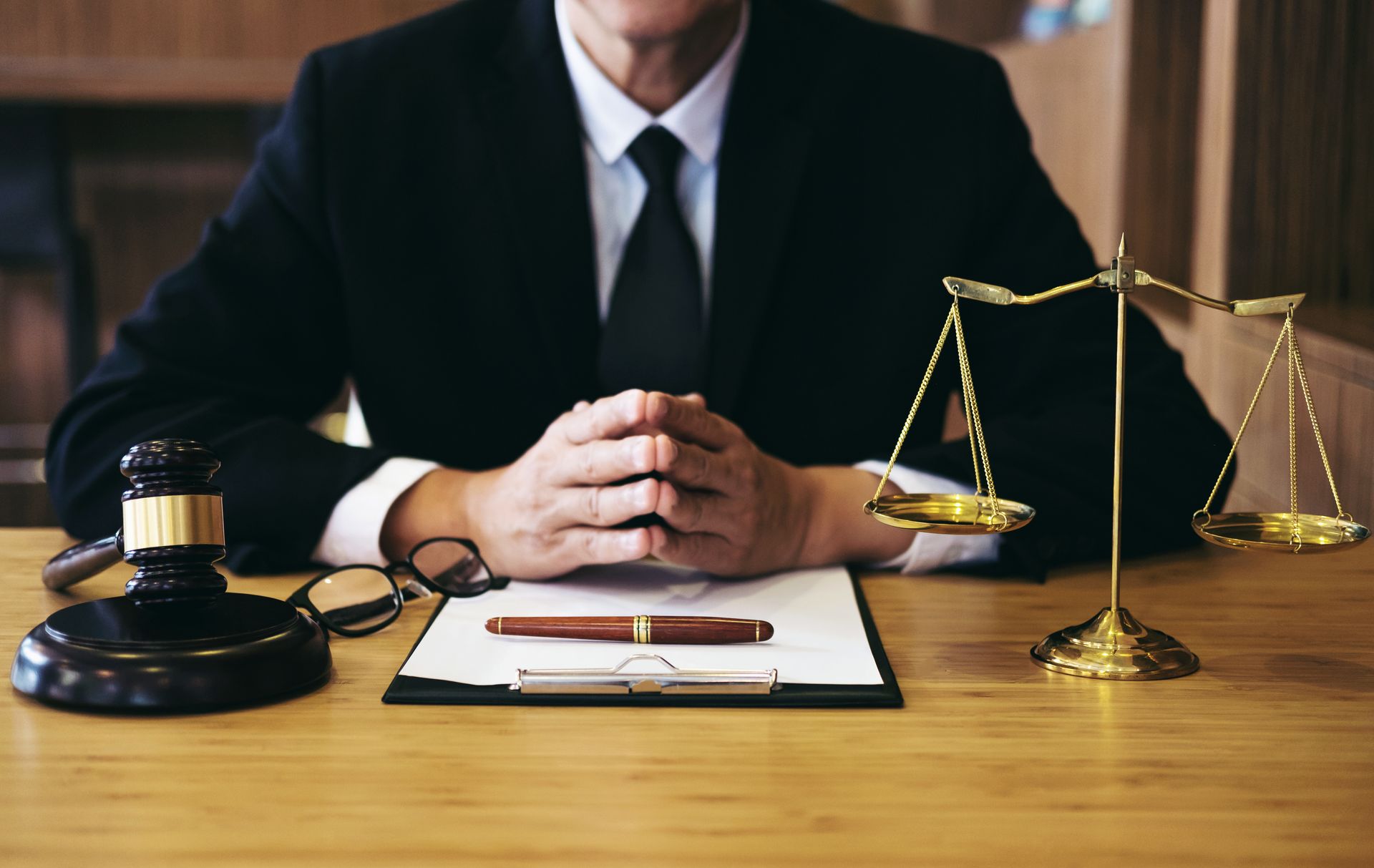 A lawyer in a suit sits at a desk, with a justice scale prominently displayed besides him
