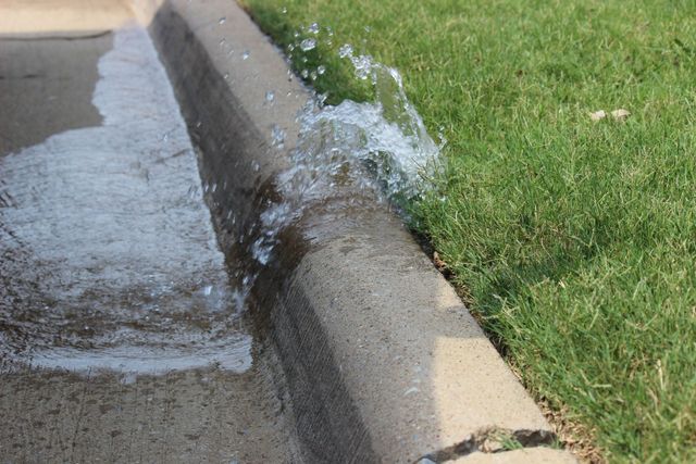 Water gushing from a curb drain onto the street beside a grassy lawn.
