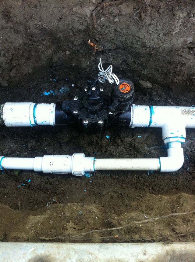 Black irrigation valve and white PVC pipes exposed in a dirt trench.