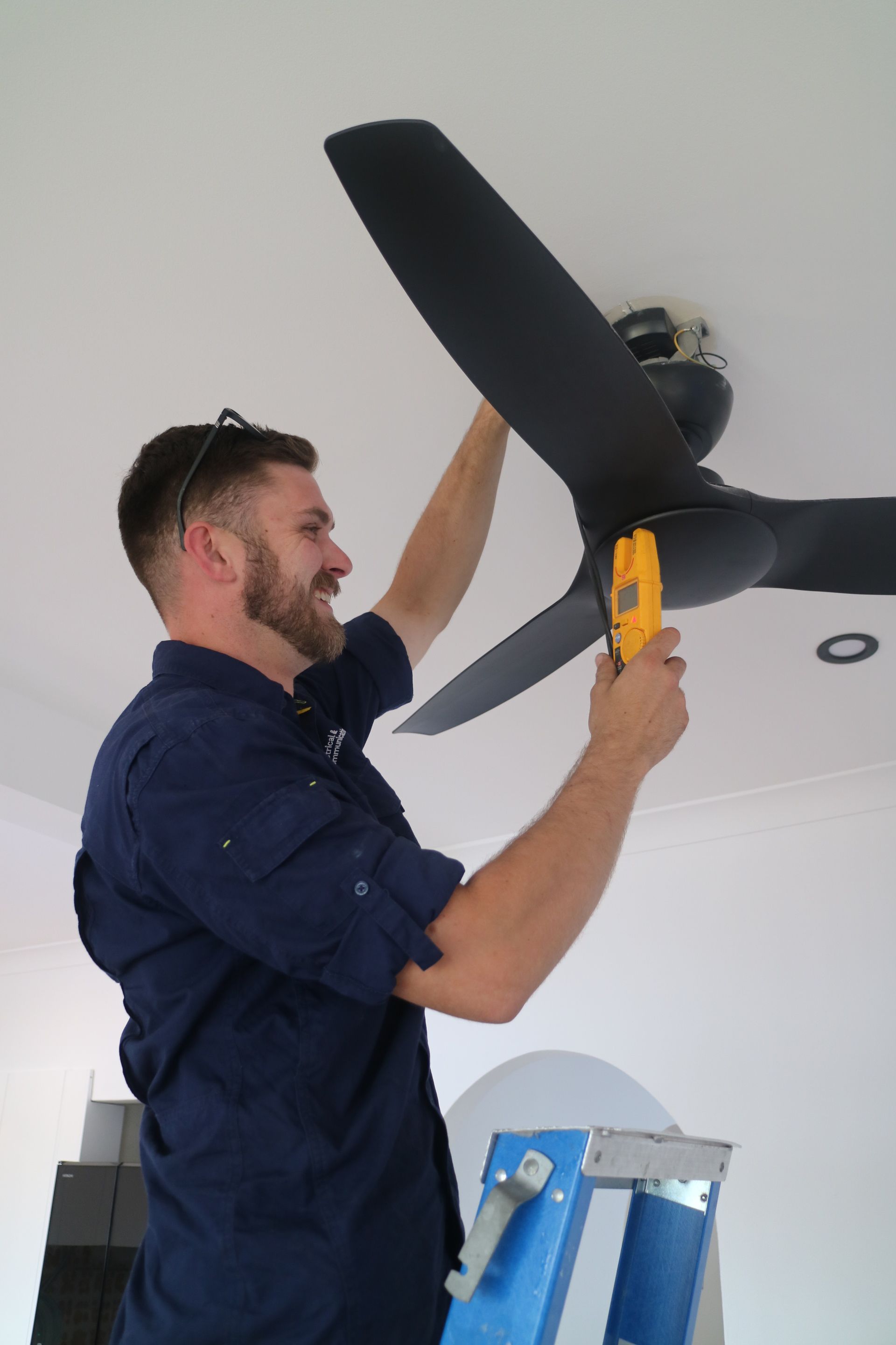 Man on Ladder Installing a Black Ceiling Fan — TB Electrical & Communications in West Gosford, NSW