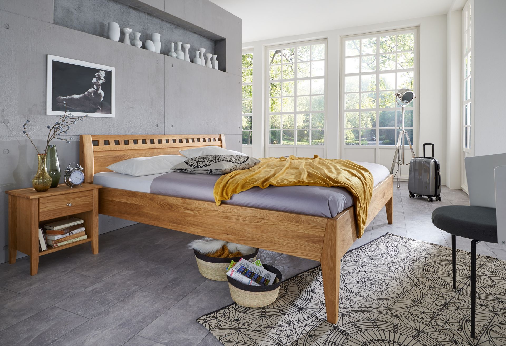 Schlafzimmer mit Holzbett, Nachttisch und grauen Wänden. Natürliches Licht strömt durch große Fenster.