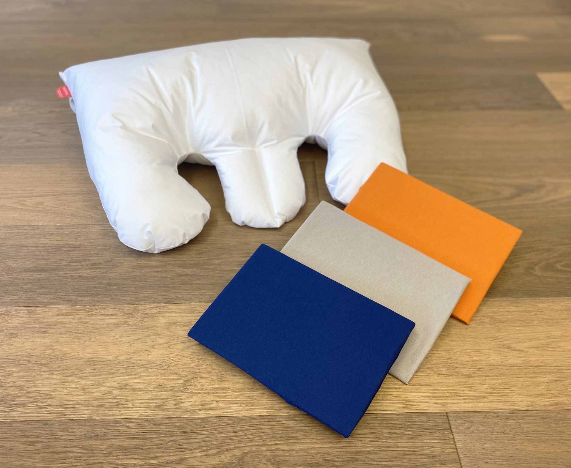 Weißes Kissen mit Gesichtsausschnitten und dreifarbigen Schaumstoffblöcken: Blau, Beige und Orange, auf einem Holzboden.