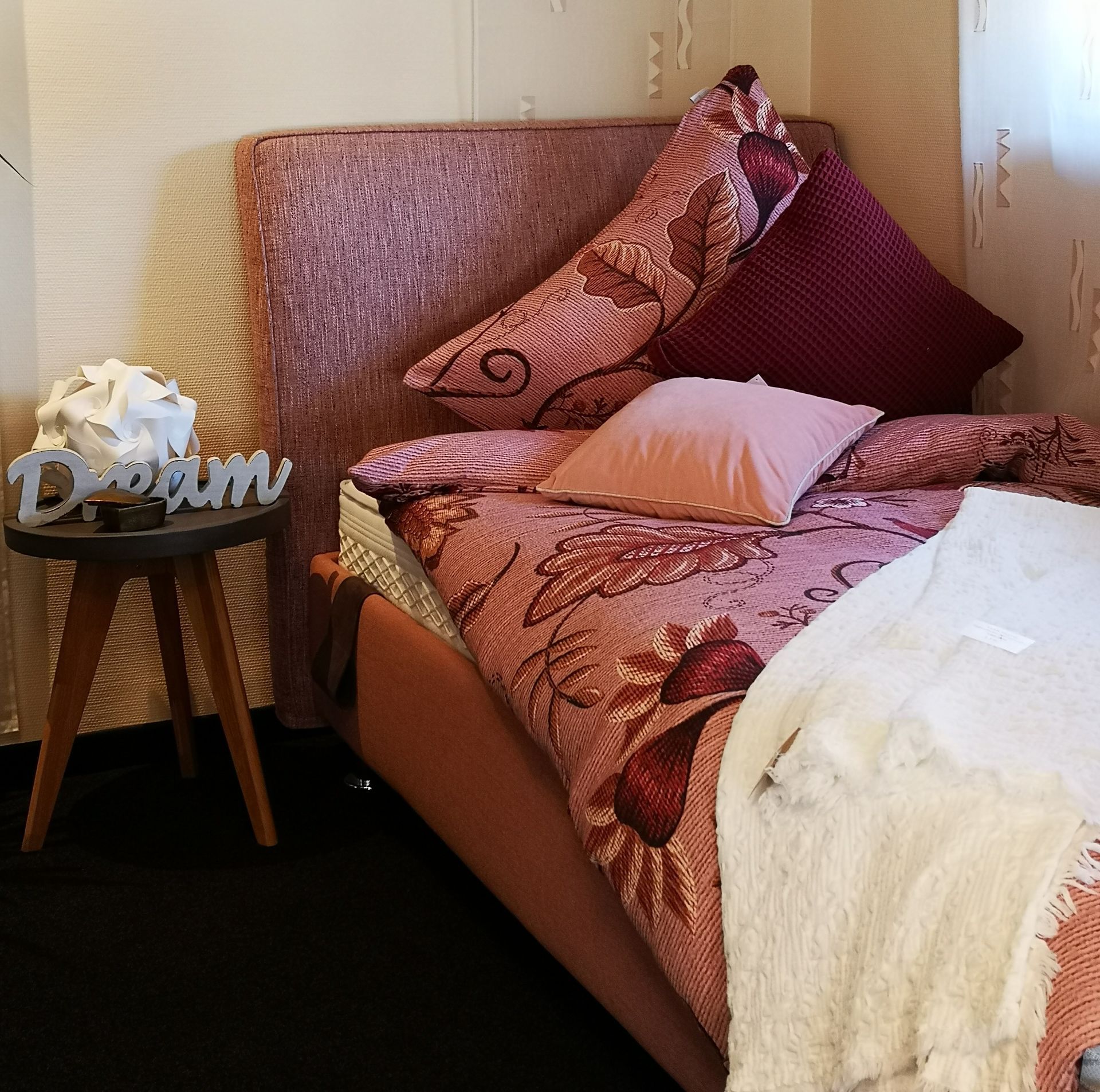 Ein Bett mit rosa geblümter Bettwäsche, einer Kunstpelzdecke und einem dekorativen „Dream“-Schild auf einem Beistelltisch.