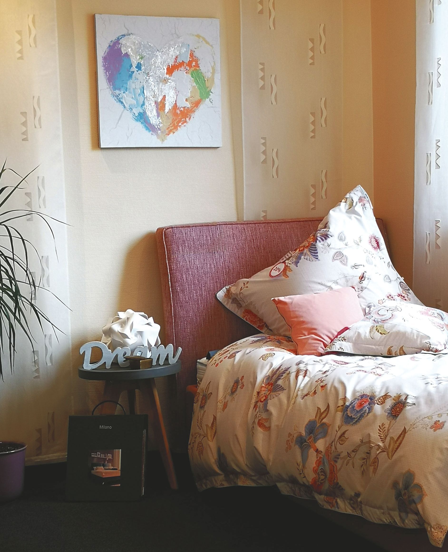 Eine gemütliche Schlafzimmerecke mit einem geblümten Bett, einem rosa Kopfteil und einem herzförmigen Kunstwerk darüber. 