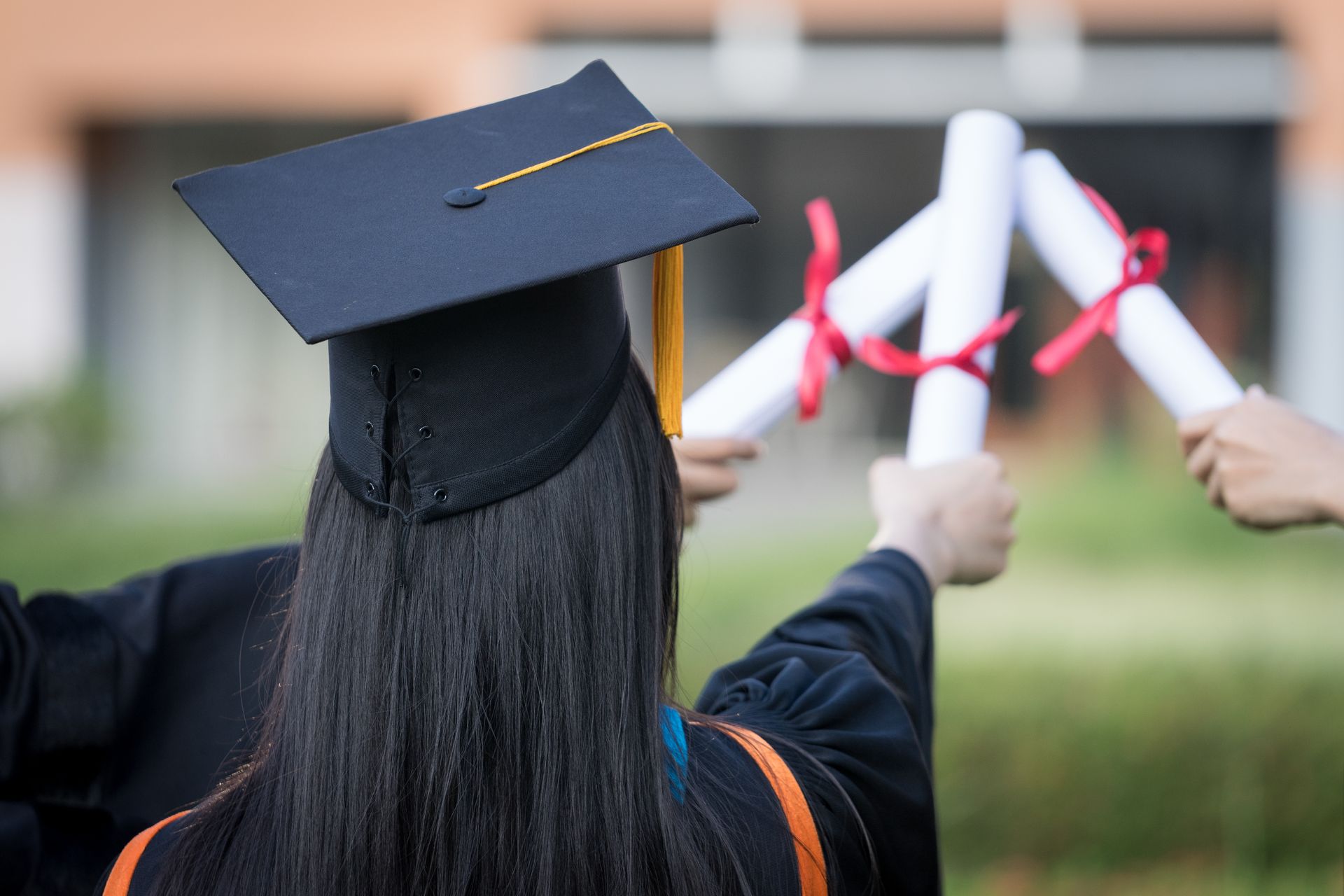 A graduate wearing a cap holds up a diploma alongside others, celebrating a graduation ceremony outdoors.