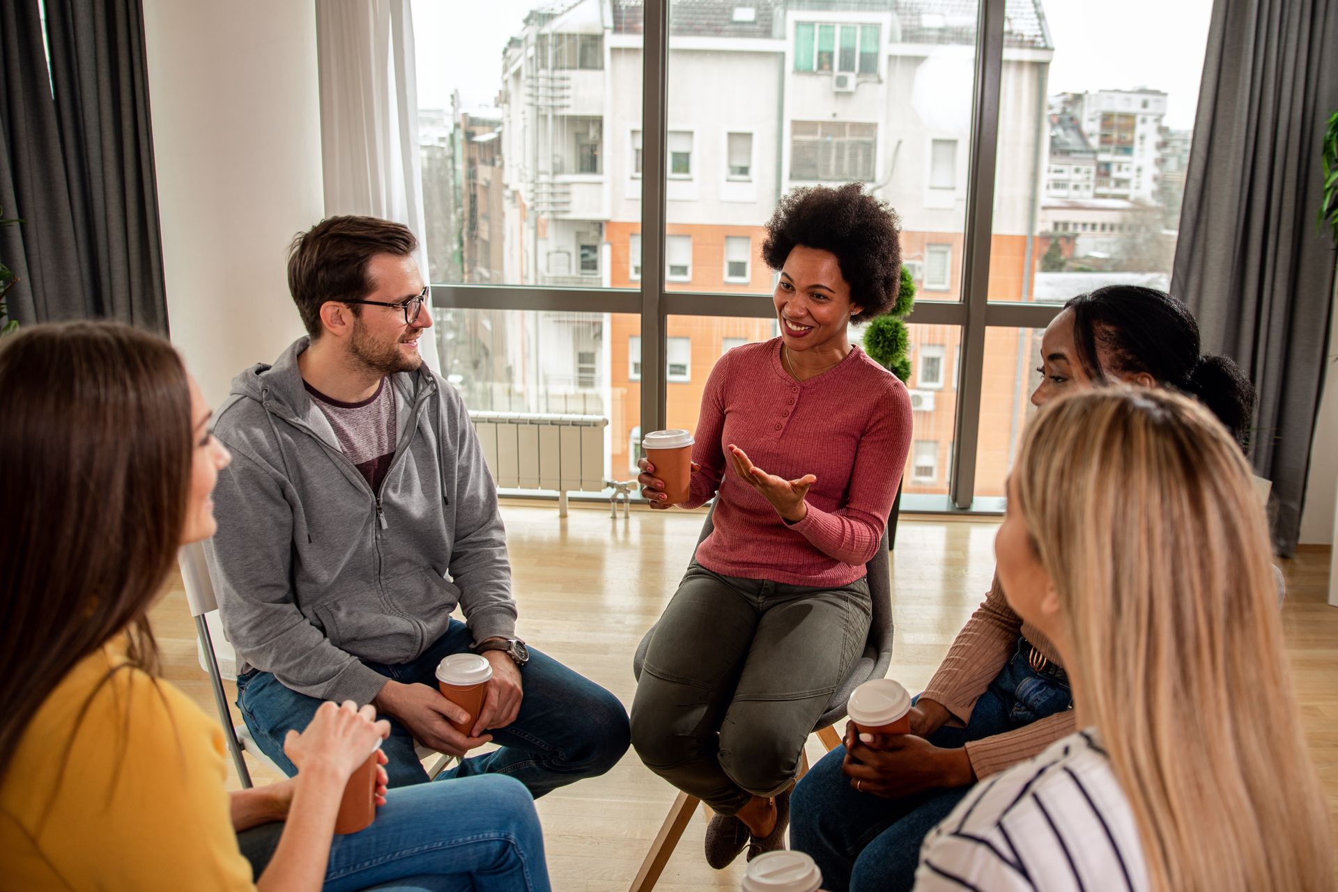 A group of people sit in a circle in a bright room, holding paper cups and talking during a casual gathering.