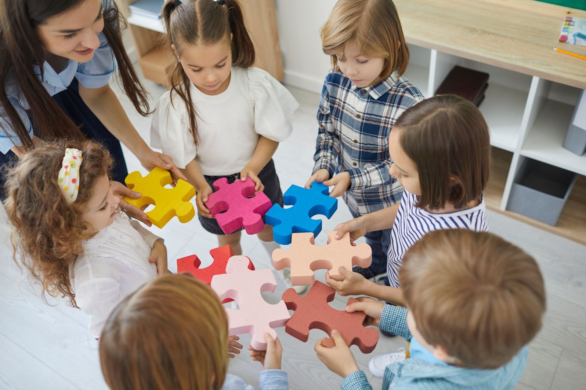 A group of children and a teacher stand in a circle, holding and connecting colorful puzzle pieces on the floor.