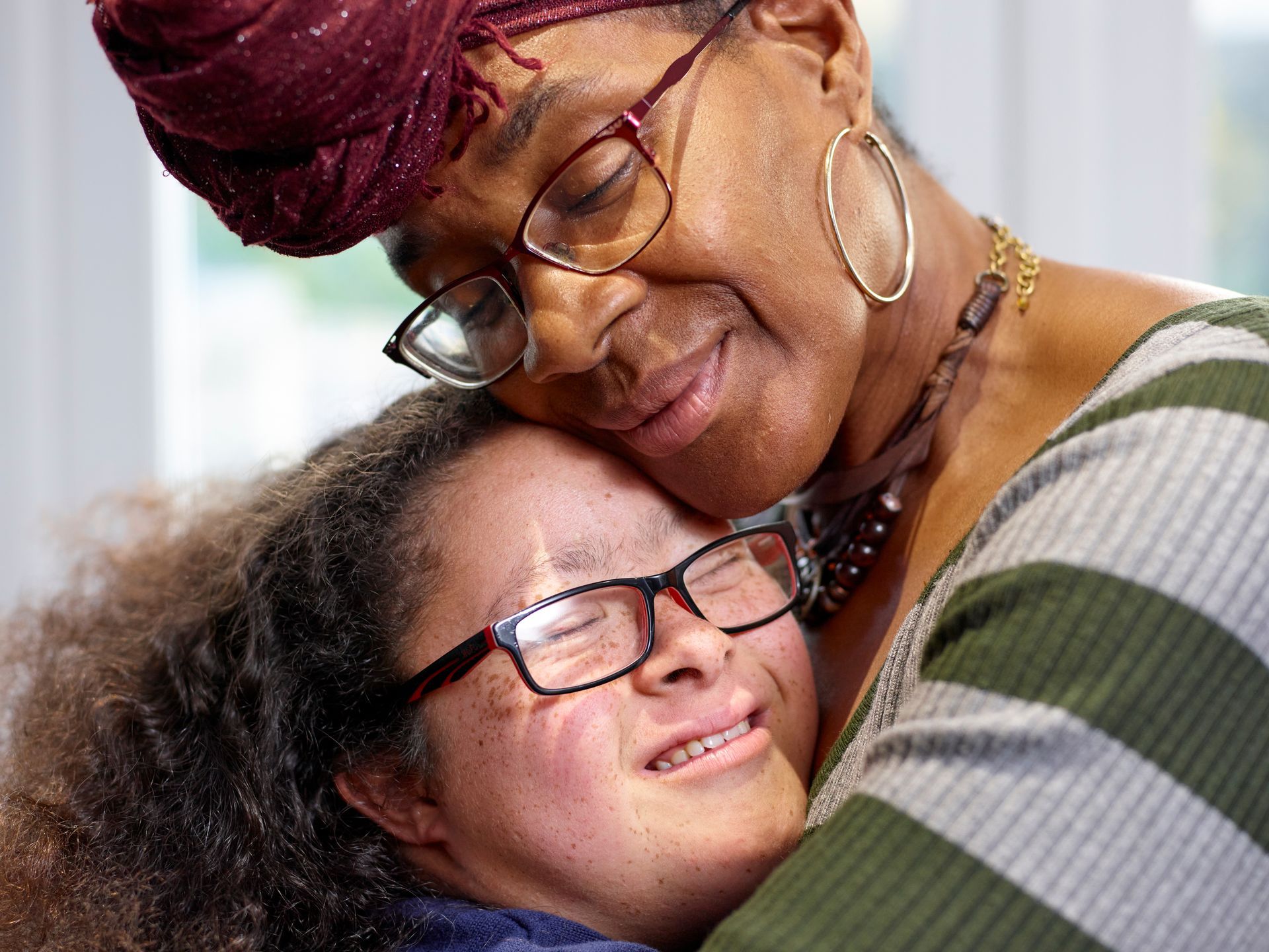 An older person and a younger person embrace closely, both wearing glasses and expressing warmth.