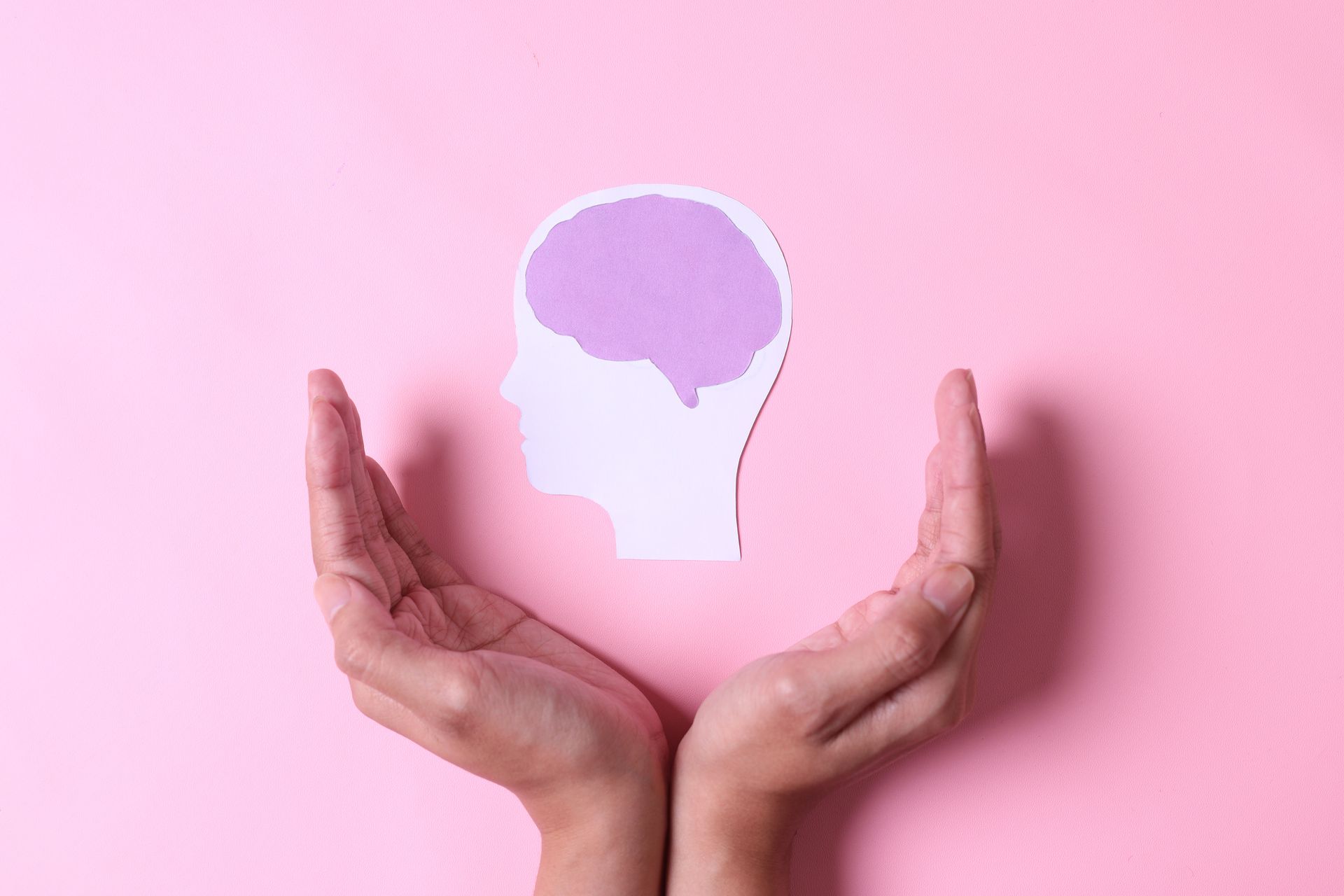 Two hands cradling a paper cutout of a human head containing a purple brain on a pink background.