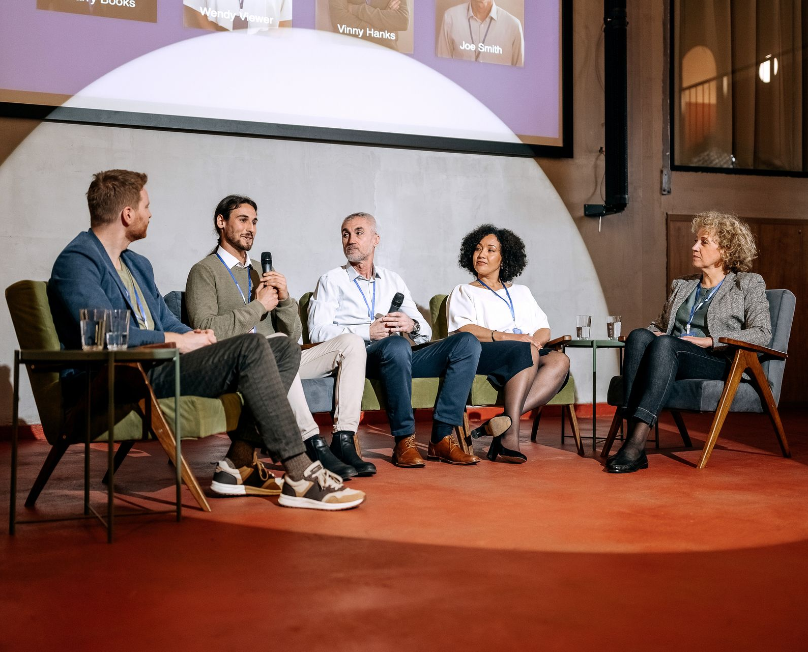A panel of four professionals sits on chairs and a sofa on a stage, discussing in a brightly lit, modern conference space.