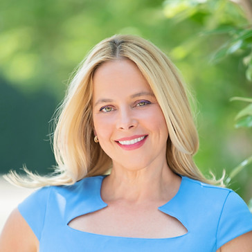 A smiling blonde woman in a blue dress with a cutout neckline, posed outdoors against a soft-focus green background.
