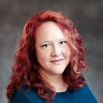 A person with shoulder-length, curly red hair looks at the camera with a neutral expression against a gray background.