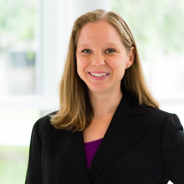 A person smiling, wearing a black blazer over a purple top, set against a blurred, bright background.