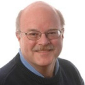 A smiling person with glasses, a mustache, and a bald head wearing a blue collared shirt under a dark sweater.
