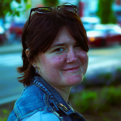 A person with dark hair and sunglasses on their head smiles at the camera, wearing a denim jacket outdoors.