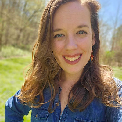 A smiling woman with wavy light brown hair and a denim shirt outdoors in a grassy, sunlit field.