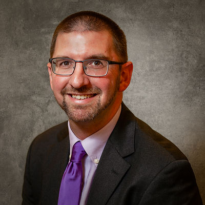 A smiling man wearing glasses, a dark suit jacket, a light-colored shirt, and a bright purple necktie.