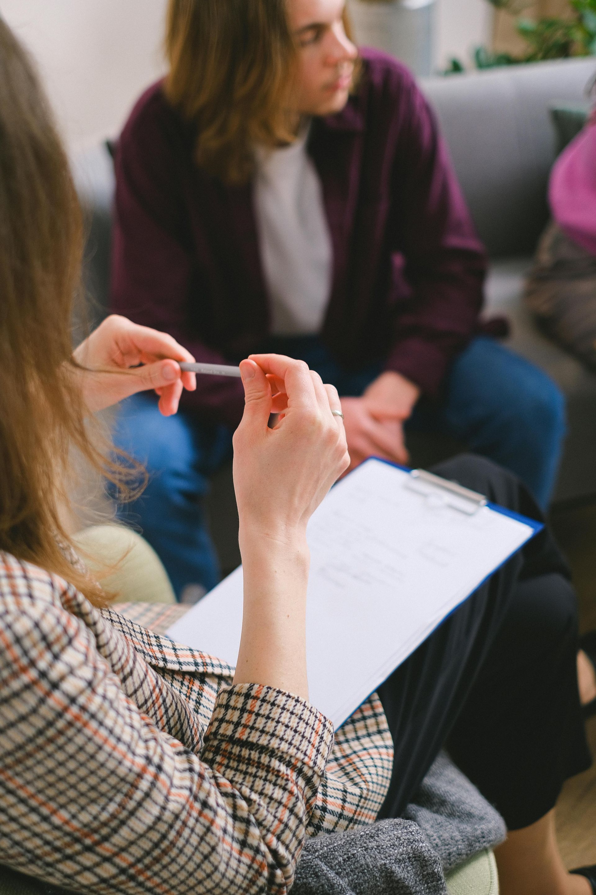 A professional holds a clipboard and pen while sitting across from a person in an office setting.