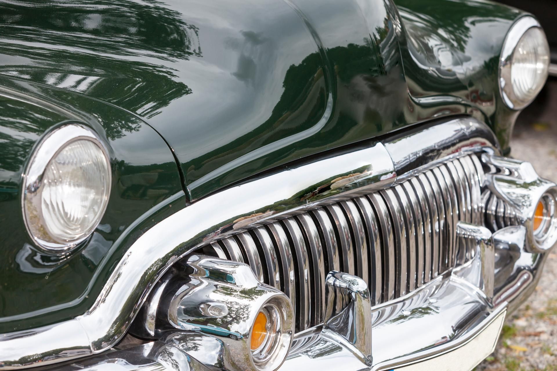 Close-up of a dark green vintage car’s chrome grille and headlight.