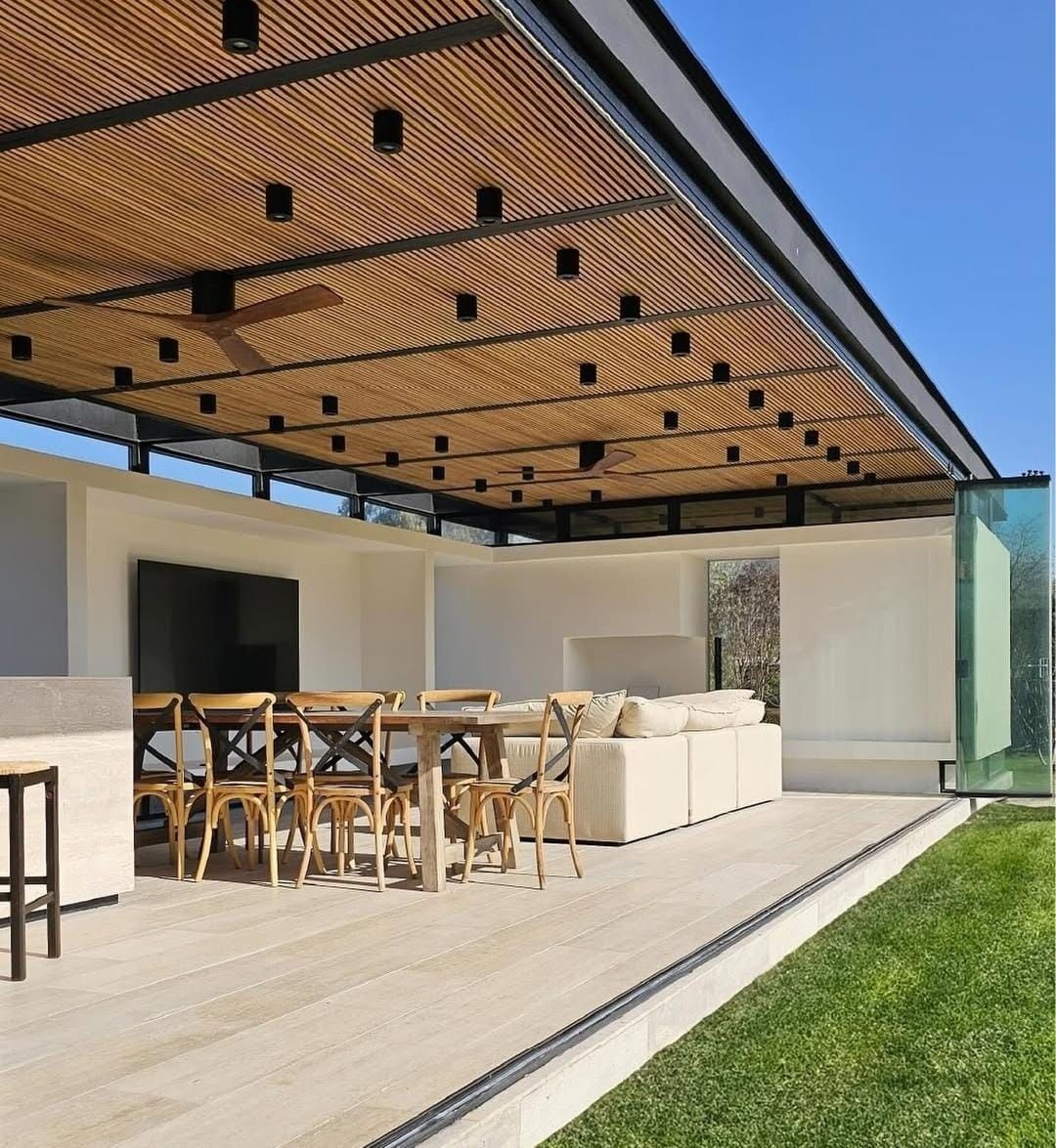 Patio moderno con pérgola, mesa de comedor, sofá beige y césped verde junto a una casa blanca.