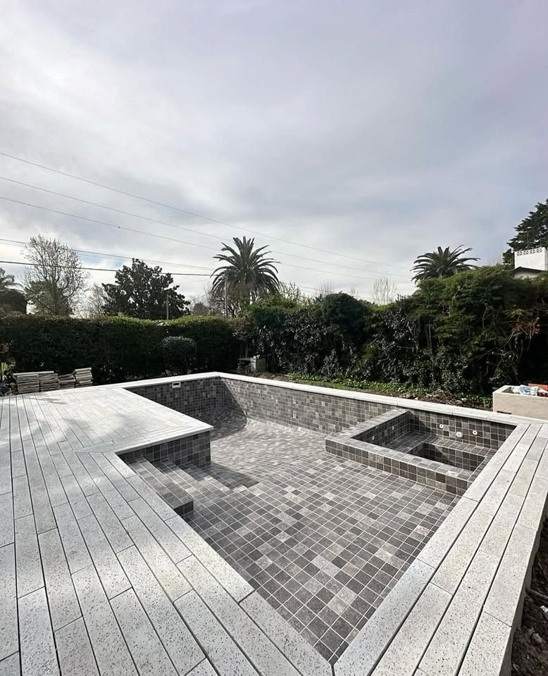 Piscina vacía en el patio trasero, con azulejos, borde gris y setos a su alrededor, bajo un cielo nublado.