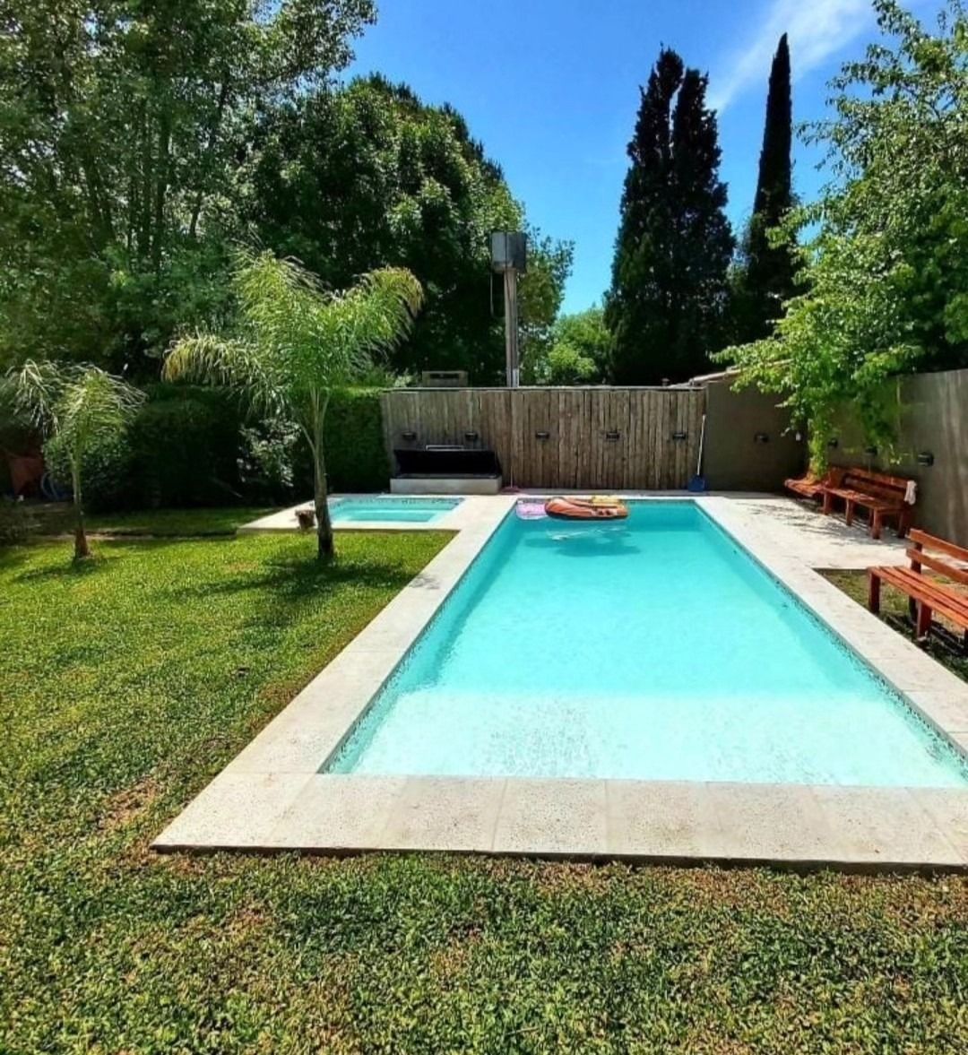 Patio trasero con una estrecha piscina turquesa, césped, árboles y valla de madera bajo un sol brillante.