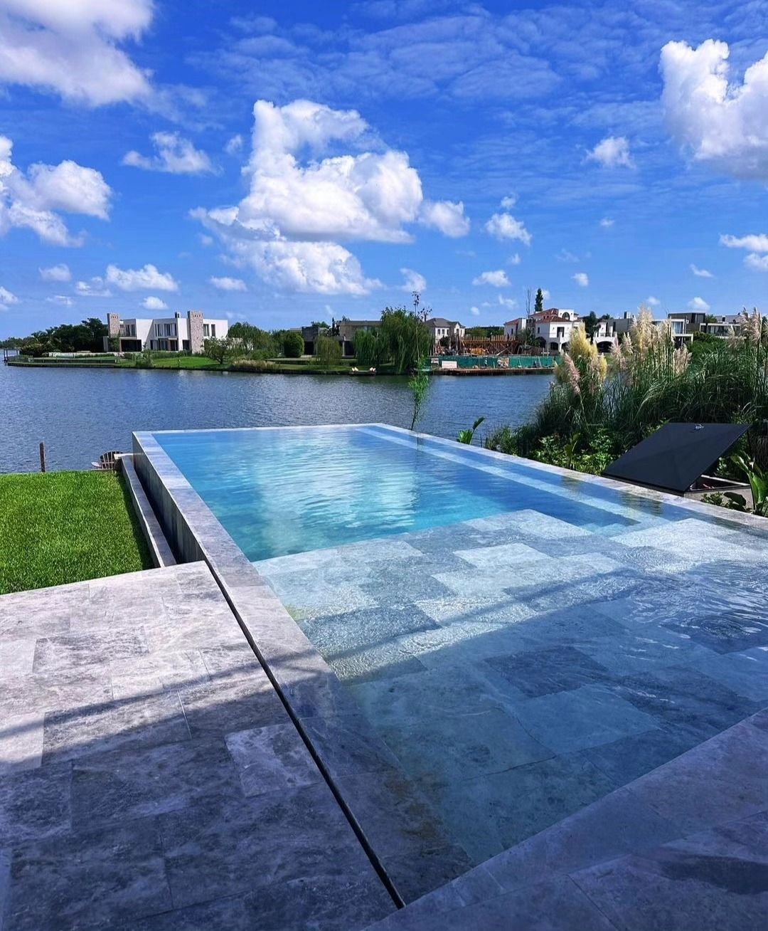 Piscina infinita con vistas a un lago y edificios lejanos bajo un cielo azul brillante con nubes.