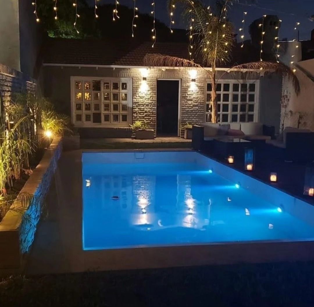 Patio nocturno con piscina azul iluminada, guirnaldas de luces y una fachada de casa moderna.