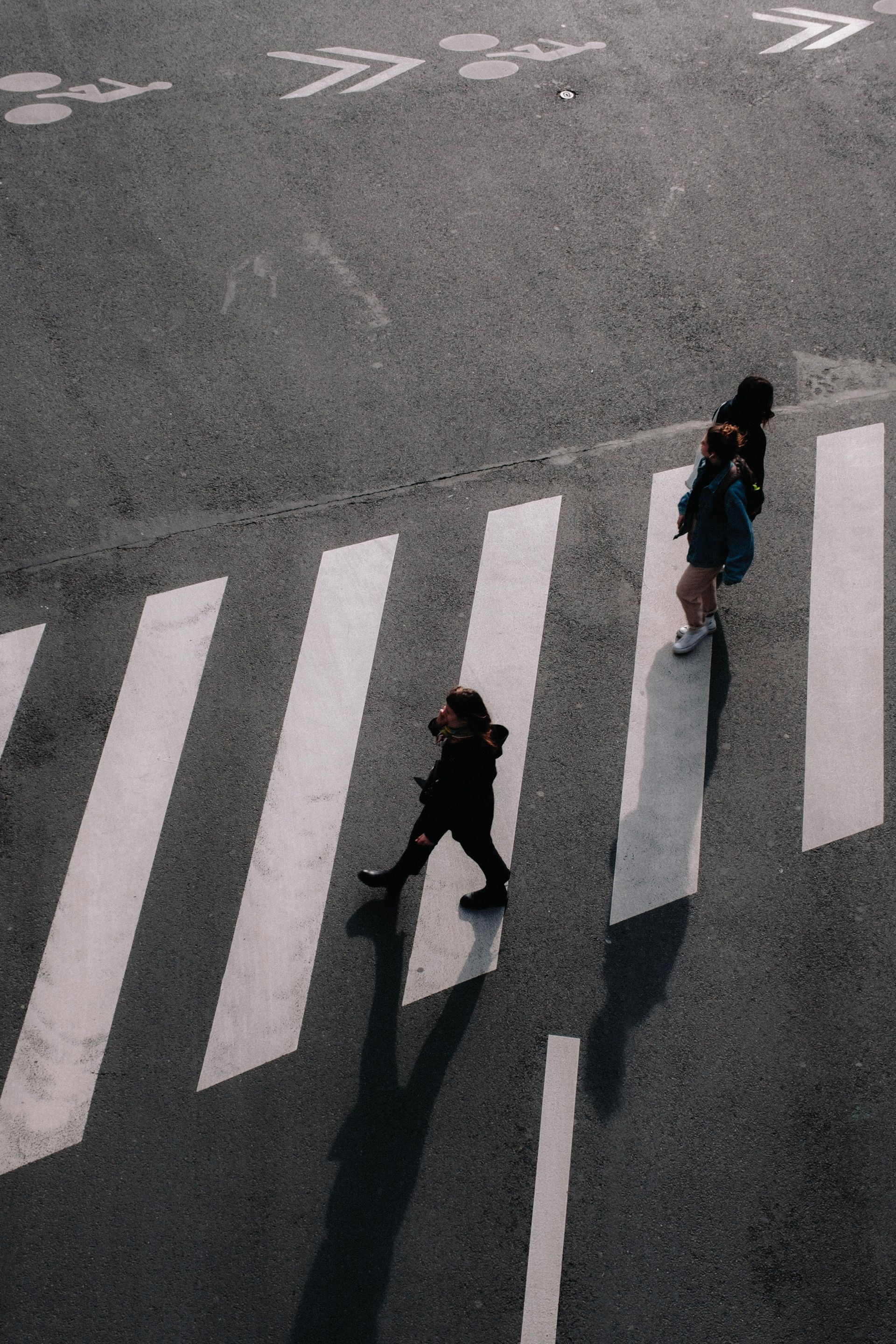 Two people crossing a wide crosswalk on a gray street, with long shadows cast by the sun.