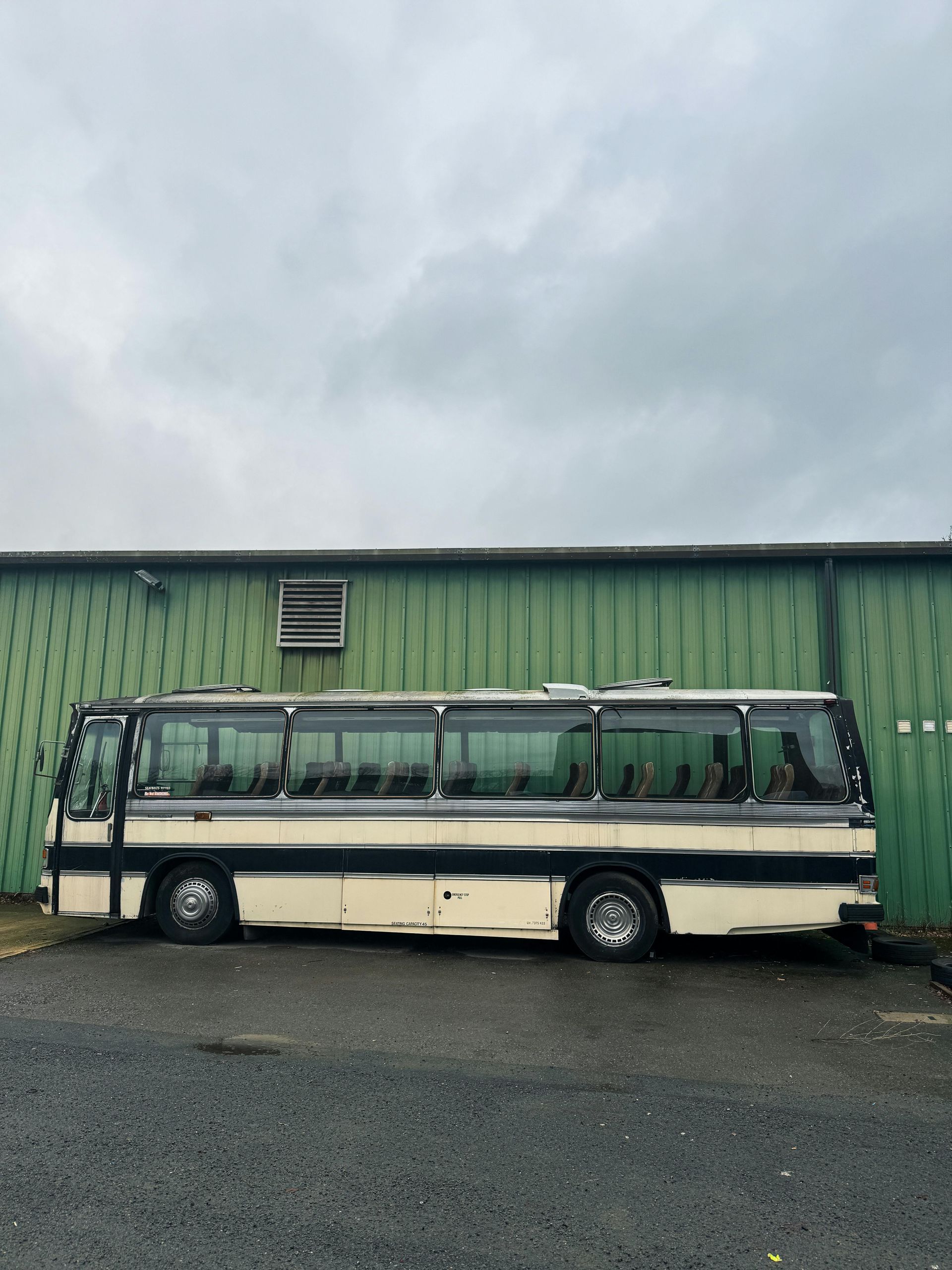Bus parked against a green building under a cloudy sky. The bus is cream and black.