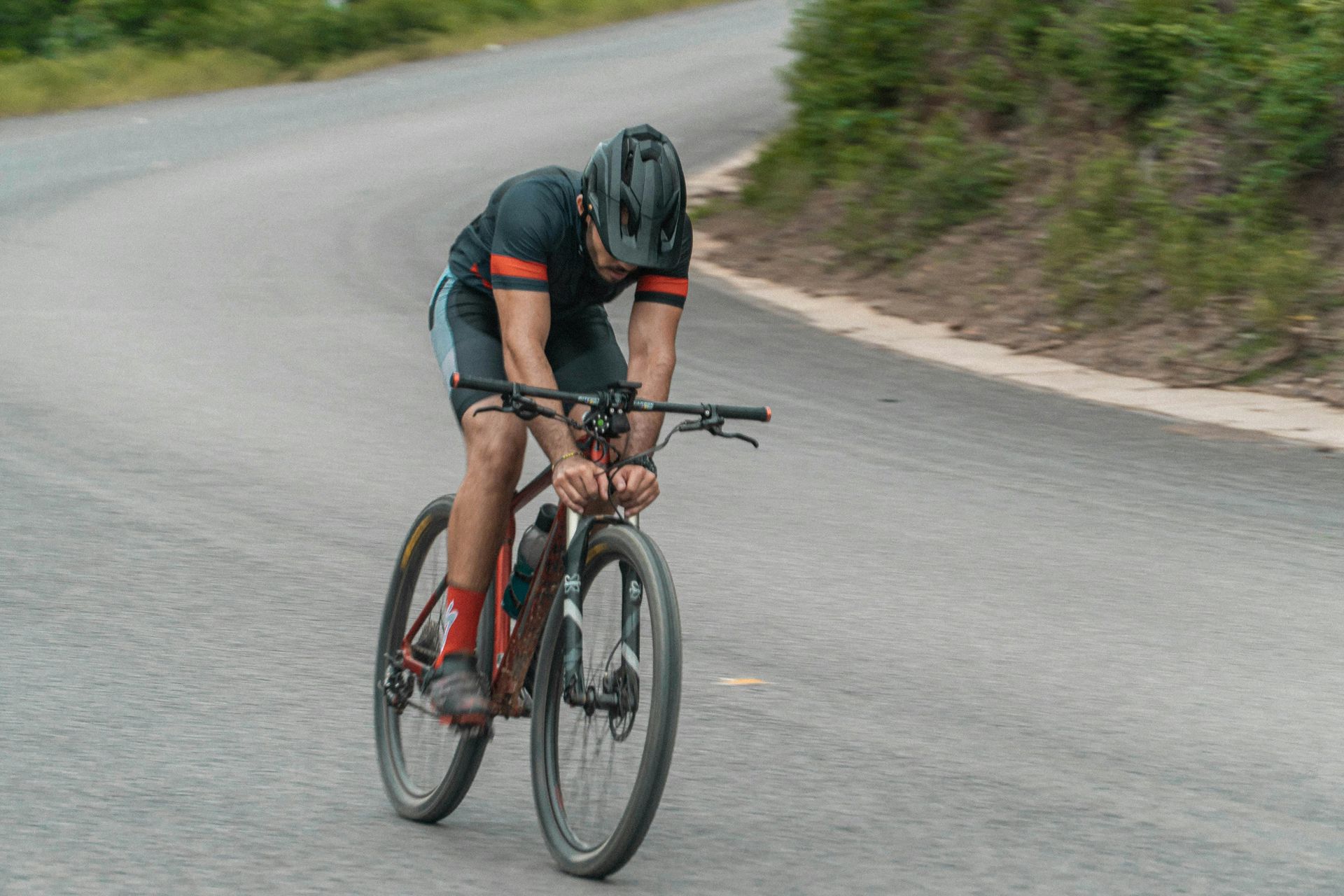 Cyclist in a black helmet and jersey leans over handlebars on a paved road, riding a mountain bike.