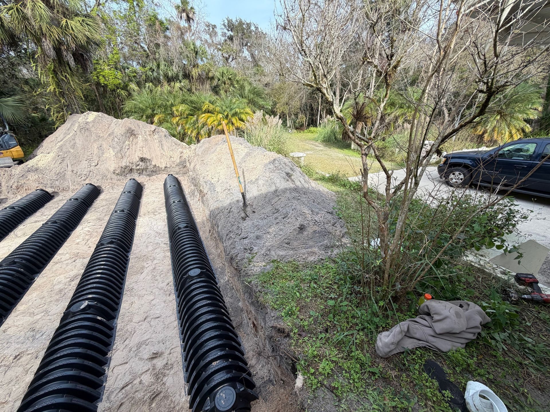 Black drainage pipes in sand, beside a driveway. Pile of sand and brush in the background. Sunny day.