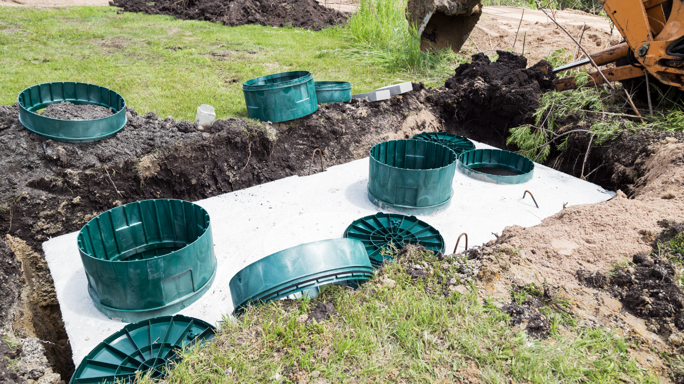 Installation of a septic system: green tanks, white base, in a dirt trench, heavy machinery in the background.
