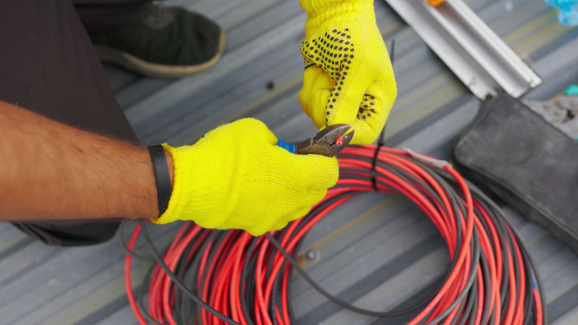 Person using pliers to cut a black wire, wearing yellow work gloves, near a coil of red wires.