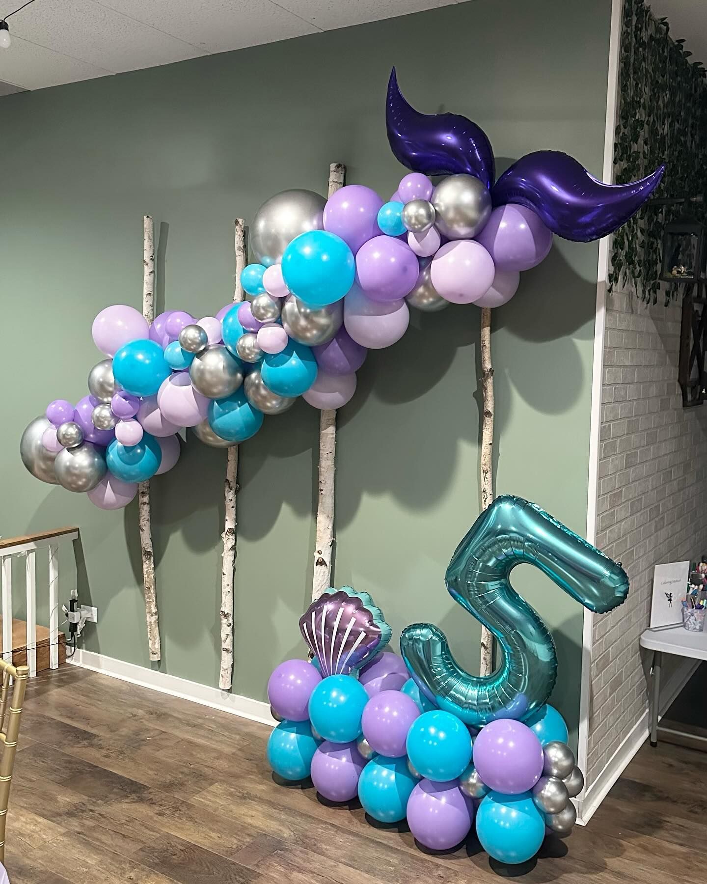 Purple, teal, and silver balloon garlands decorate a wall with a large teal balloon number 5.