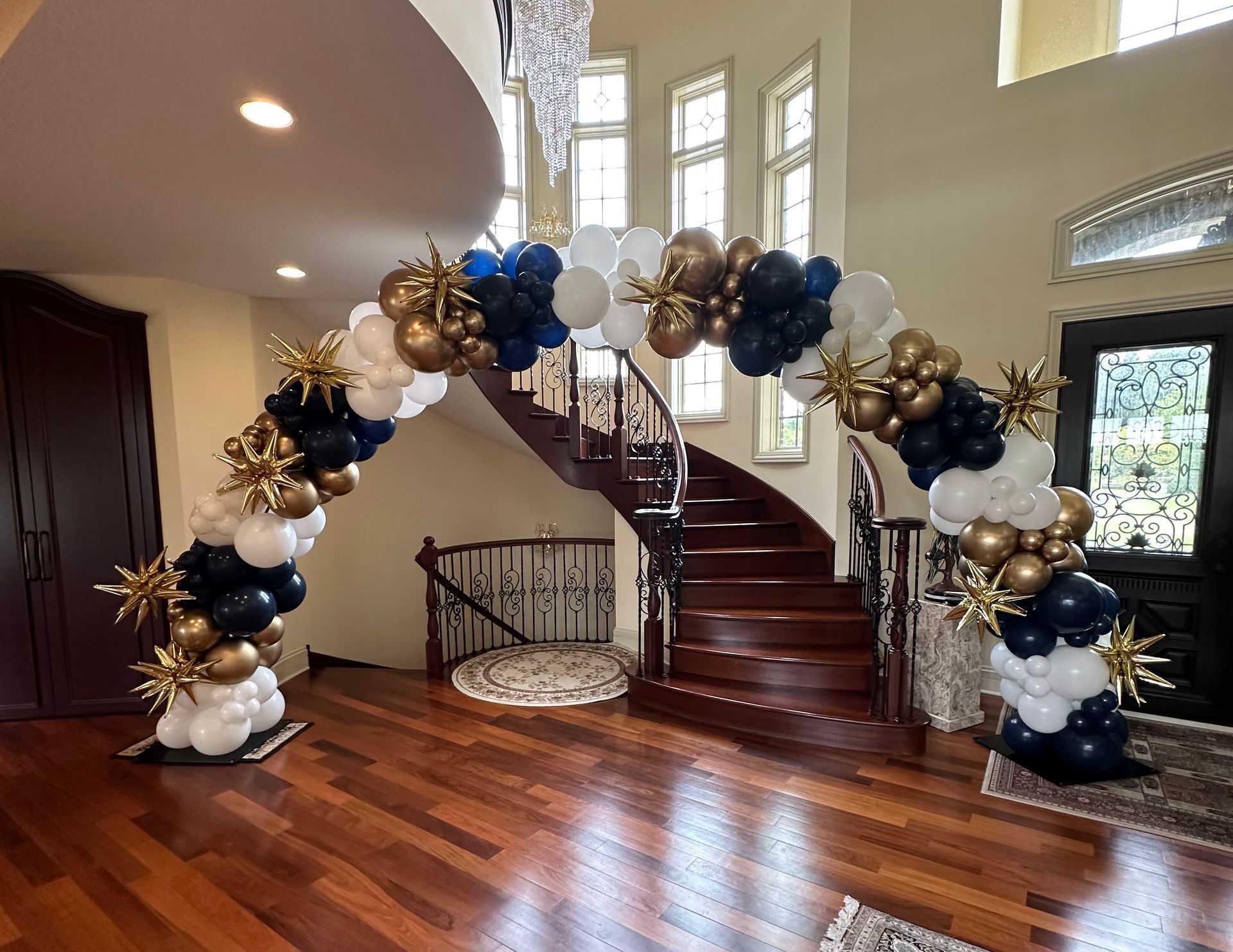 Black, gold, and white balloon arch in a foyer with a curved staircase and hardwood floors