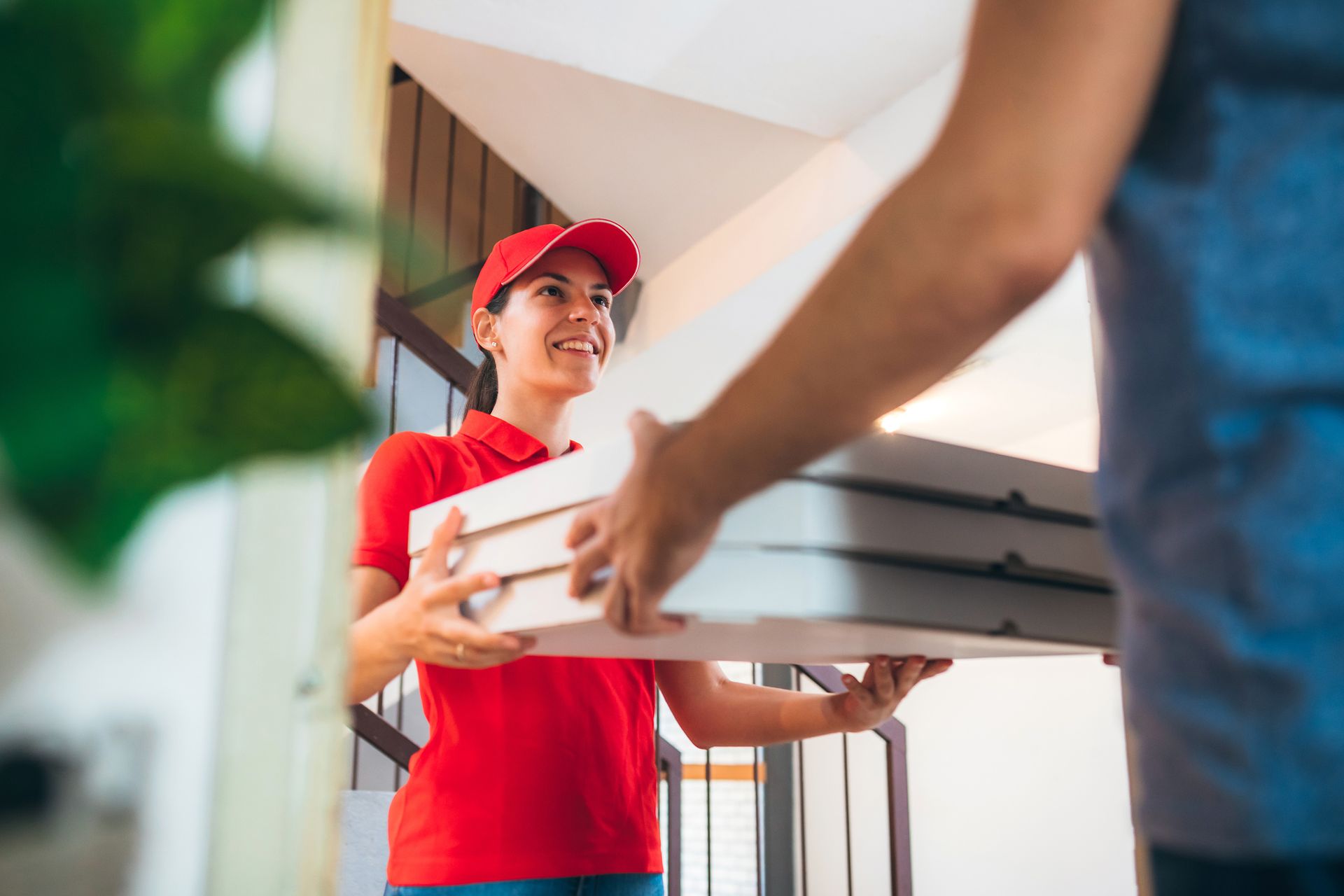 A pizza delivery woman wearing a red cap and t-shirt giving pizza boxes to a customer.