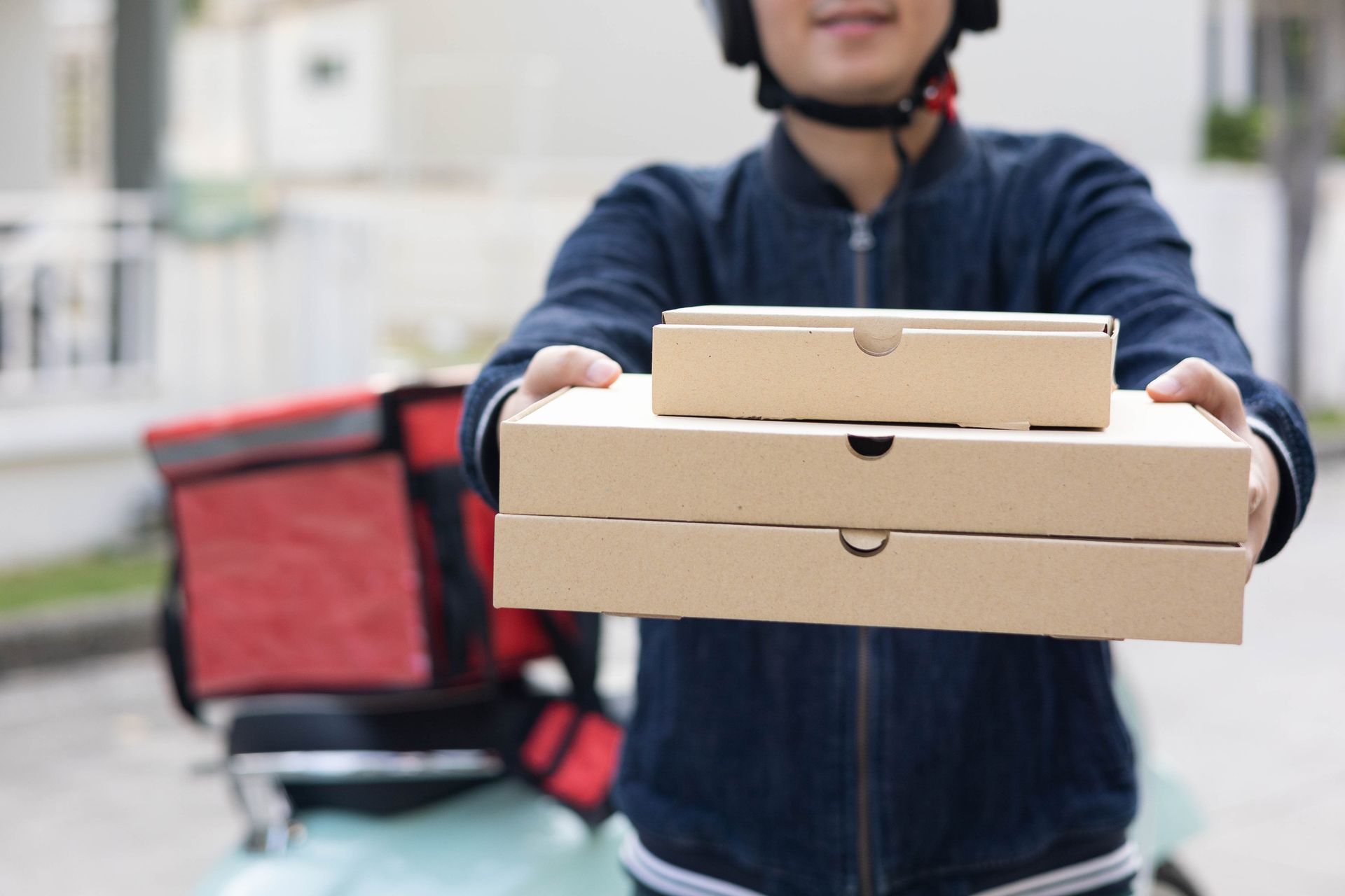 Delivery person holding pizza boxes with red delivery bag and scooter in urban background. Delivery person holding pizza boxes with red delivery bag and scooter in urban background.