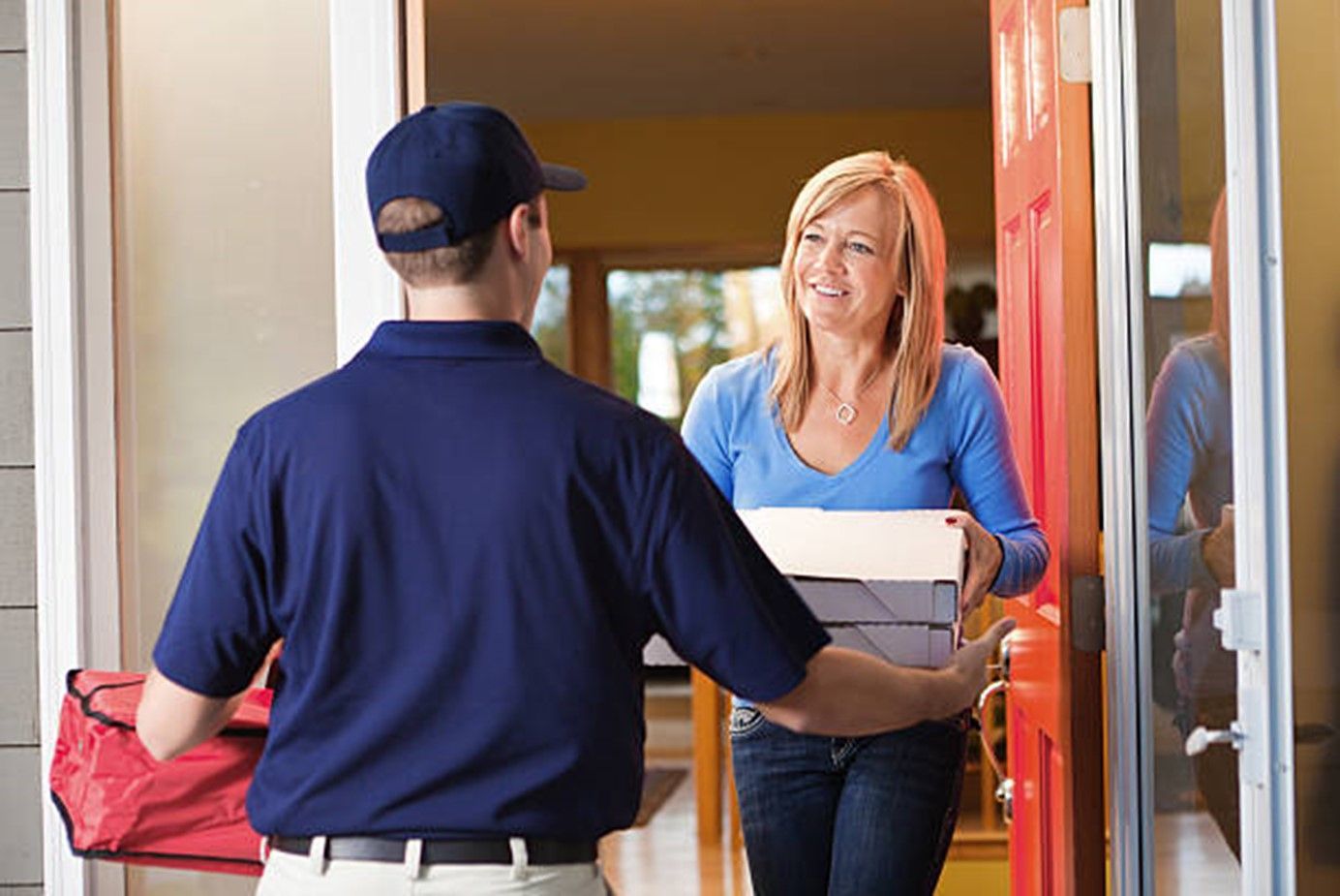 Pizza delivery driver in navy uniform hands white pizza boxes to woman at orange front door. 